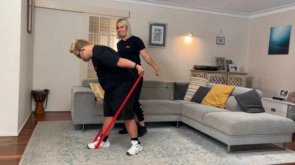 A coach alongside a young man doing a theraband exercise in a living room, from an Able video.